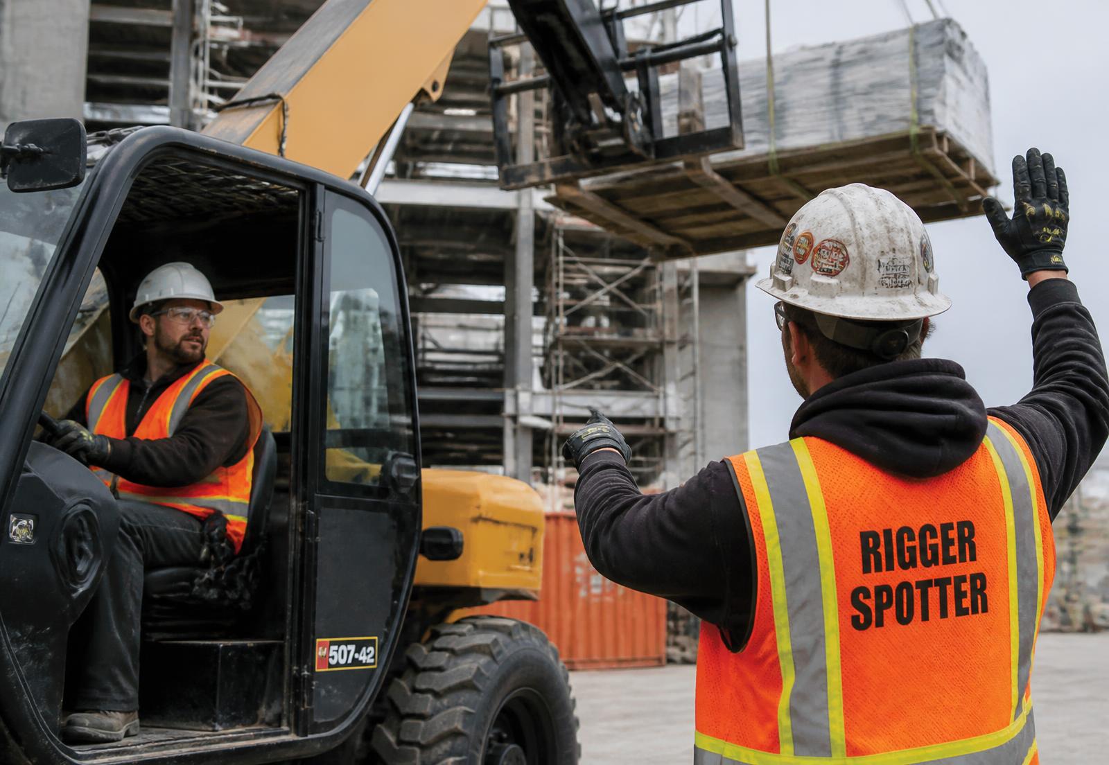 Rigger in a high-visibility vest and hard hat signaling a telehandler operator on a construction site